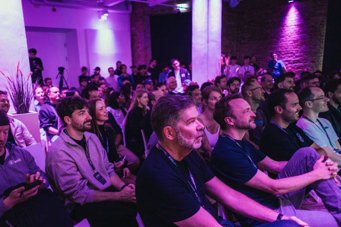 The image shows a large group of people sitting down, possibly at an event or gathering. They are all wearing dark clothing, which suggests that they might be attending a formal or semi-formal occasion. The attendees appear to be engaged in what appears to be a presentation or discussion, as some individuals have their hands on their laps and others are looking attentively towards the speaker.

The room is filled with people sitting in rows of chairs, creating an atmosphere of concentration and interest. There are also potted plants placed throughout the space, adding a touch of greenery to the scene. The image captures the essence of a formal event or gathering where attendees are focused on listening and participating in the discussion.