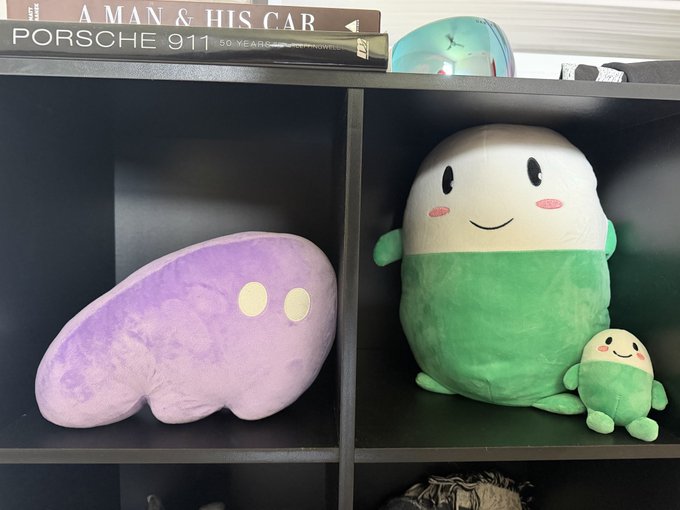 A purple Phantom (PHANTOM) crypto plushie sits on a shelf next to a green and white Cupsey plushie, with books titled "A Man & His Car" and "Porsche 911" on the top shelf. The image illustrates the post's message by showcasing two popular crypto-themed plushies representing their respective communities, supporting the claim that $CUPSEY will reach a $100M market cap.