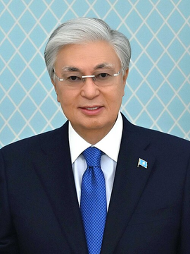 A formal portrait of Kazakhstan's President Kassym-Jomart Tokayev, dressed in a dark suit with a blue tie and a Kazakhstan flag lapel pin, against a light blue patterned backdrop. The image accompanies news of his decree advancing the country's digital asset industry with crypto-friendly reforms.