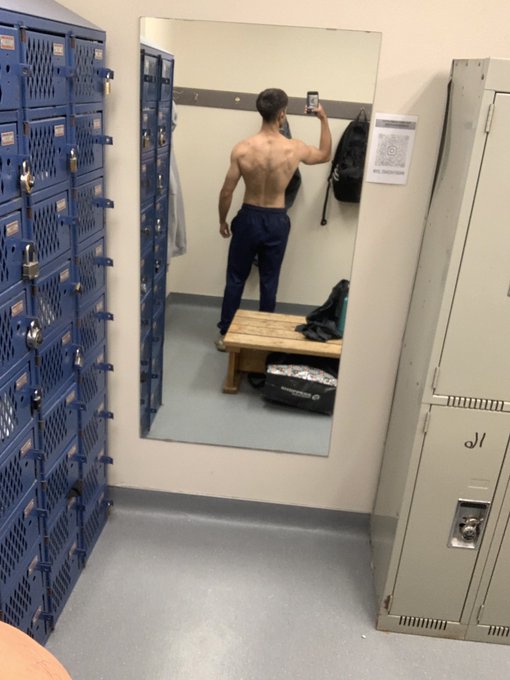 A mirror reflection in a locker room shows a shirtless man taking a photo with his phone, standing on a bench next to blue lockers, with a backpack on the floor.
