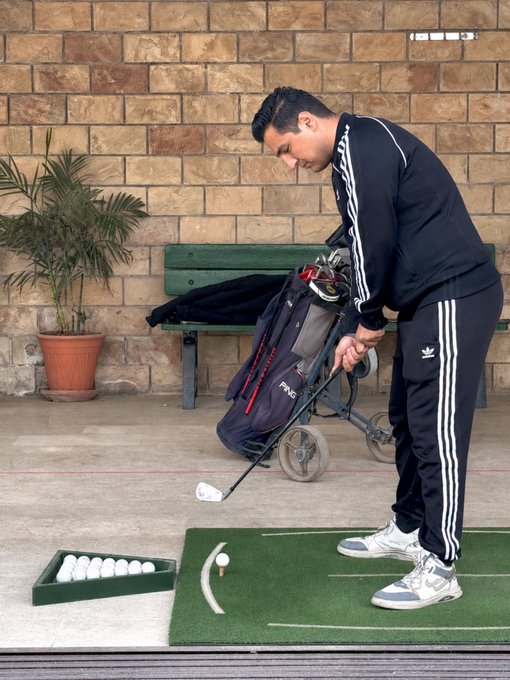 The image shows a man wearing black pants, a black jacket with white stripes, and white shoes standing next to a golf cart that has several golf balls scattered around it. The man appears to be practicing his swing at a driving range or preparing for a game of golf. The post on the wall reads "Good Morning Everyone Who will say 'GM BACK'?". This suggests that the image might have been taken during a morning practice session, and the man is likely getting ready for a round of golf with friends or family members who are participating in the activity.