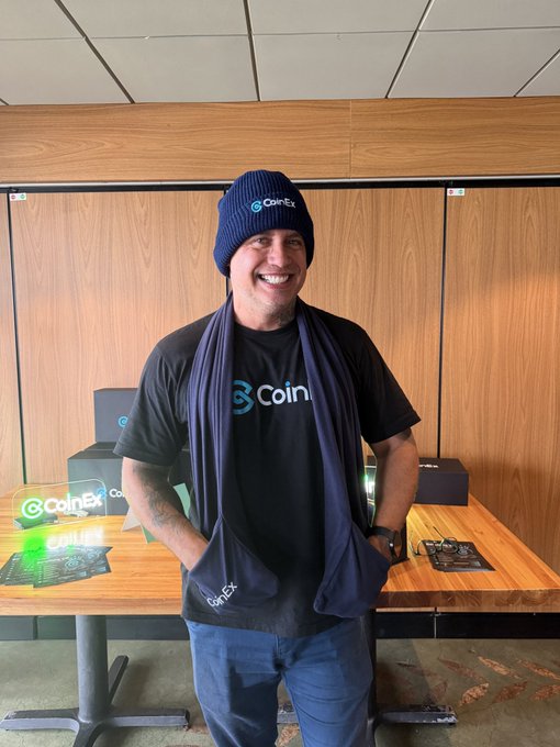 A smiling man wearing a blue 'CoinEx' beanie, a matching blue scarf, and a black 'CoinEx' t-shirt, standing in front of a wooden table displaying CoinEx branded merchandise and holographic signs.