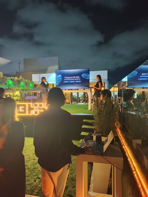 An evening outdoor tech event with a stage displaying 'Programa VIP OKX' and 'OKX' branding, featuring attendees and equipment in a dimly lit setting with decorative orange lighting.
