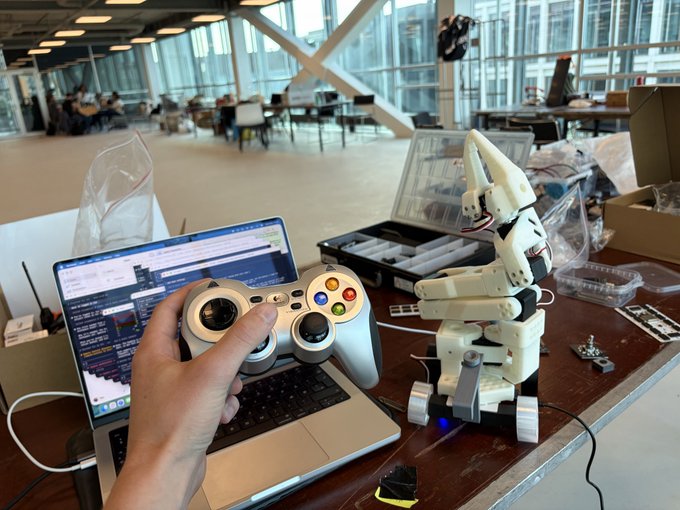 A first-person view shows a hand holding a small gamepad controller connected to a laptop, with a small 3D-printed robotic arm sitting on the desk next to it in a lab or classroom setting.