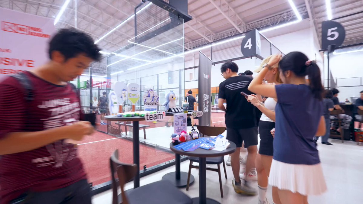 The image shows a group of people standing around a table with drinks, possibly at an event or gathering. One person is using their phone while others are engaged in conversation. The background features a large window that allows natural light into the room, creating a welcoming atmosphere for socializing and enjoying each other's company.