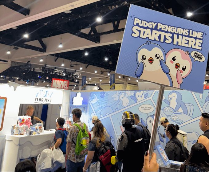 A convention floor photo showing a blue sign reading 'PUDGY PENGUINS LINE STARTS HERE' held up in front of a busy Marvel/Pudgy Penguins booth.