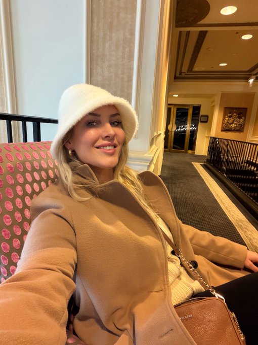 A smiling blonde woman wearing a white fuzzy bucket hat and tan coat takes a selfie in what appears to be a hotel lobby with a pink patterned chair and artwork in the background.