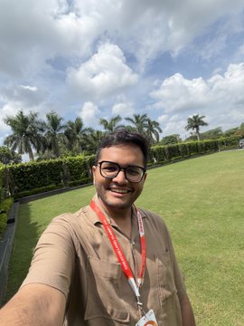 A man in a beige shirt and glasses smiles for a selfie outdoors, with palm trees and a cloudy blue sky in the background. The red lanyard around his neck suggests he is attending an event, fitting the post about Monad Blitz Bangalore v5.