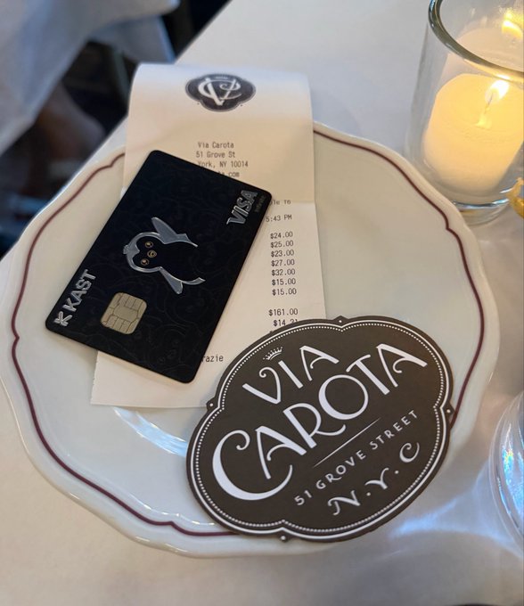 The image features a white plate with a black credit card and a receipt for a restaurant meal placed on top of it, along with a small sticker that says "via carota street" written on it. The plate has a red rim around the edge, adding a touch of color to the scene. In the background, there is another plate with a candle on it, suggesting that this might be a romantic or intimate setting for a meal.