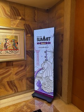 A digital signage display promotes "THE SUMMIT" with what appears to be Solana branding and event details, positioned in a hallway near a framed artwork. The image humorously underscores the post's complaint about low turnout at Solana Summit Singapore, as the event display stands alone in an otherwise empty corridor with no attendees in sight.