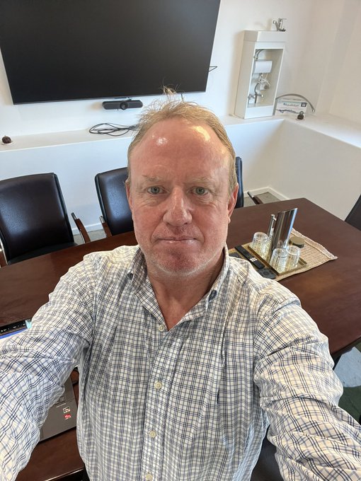 A selfie of a balding man in a checkered shirt taken in what appears to be a conference room with a large TV and table visible behind him.