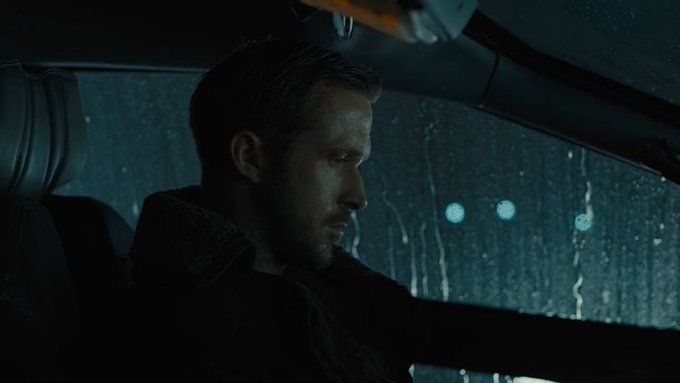 A dark, rainy car scene shows a bearded man sitting in the driver’s seat and looking downward or ahead through the windshield.