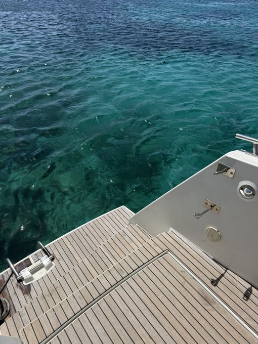 A wooden yacht deck overlooks strikingly clear turquoise water, with metal hardware and hinges visible along the white hull. The pristine teak flooring and calm sea create a sleek, ready-to-depart vibe that matches the approving "Looks good sir" tone.