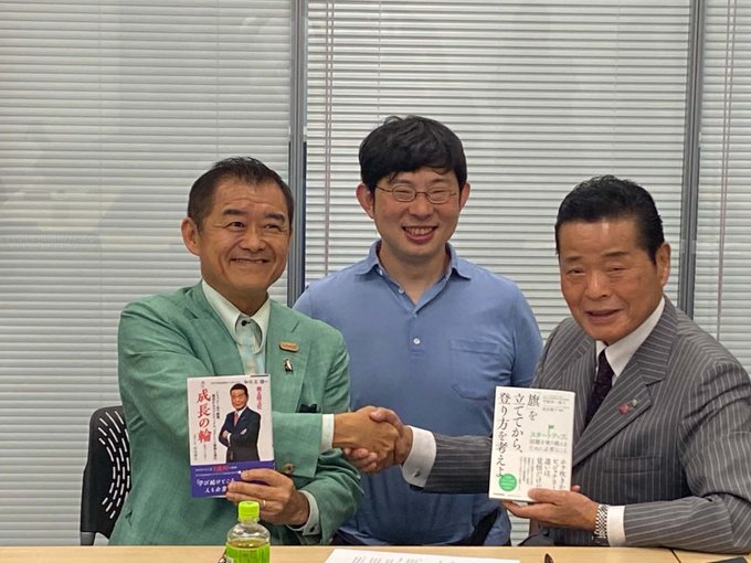 Three men in a meeting room, with two of them shaking hands while holding Japanese books; the books appear to be by 武長穂 and 齋藤元有.