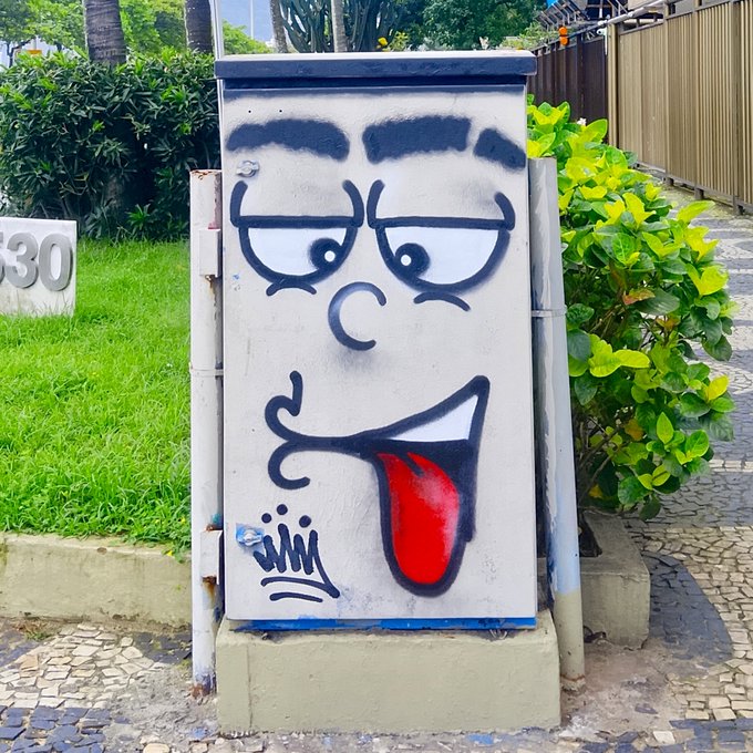 An outdoor metal utility box painted with a graffiti design of a cartoon face featuring thick black eyebrows, droopy eyes, a frowning mouth, and a red tongue sticking out to the side.