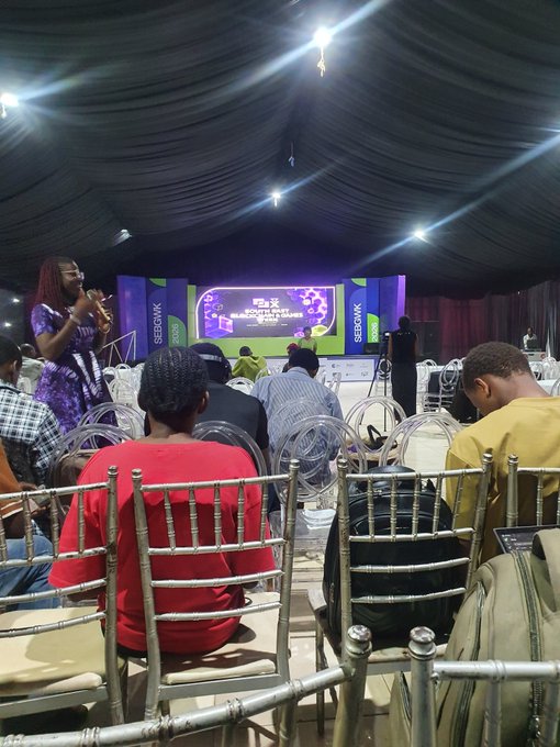An indoor event space with chairs arranged for attendees facing a stage with a screen displaying 'Segewik' branding, while a woman in purple speaks to the audience.