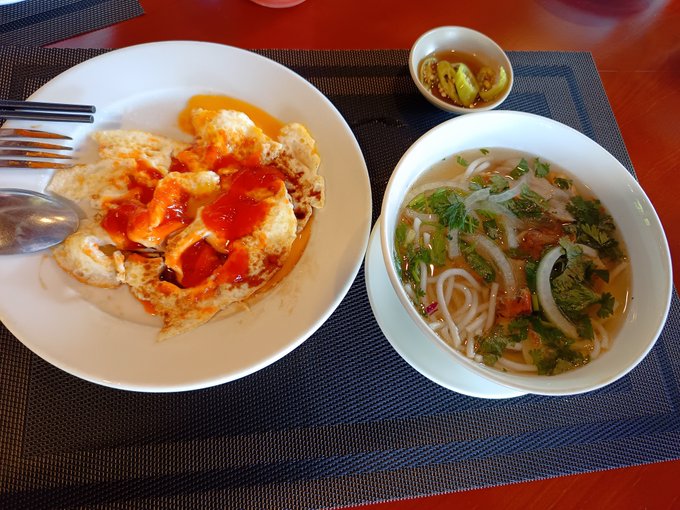 A meal consisting of a fried egg topped with red sauce on a white plate, accompanied by a bowl of noodle soup garnished with cilantro, served on a dark woven placemat.