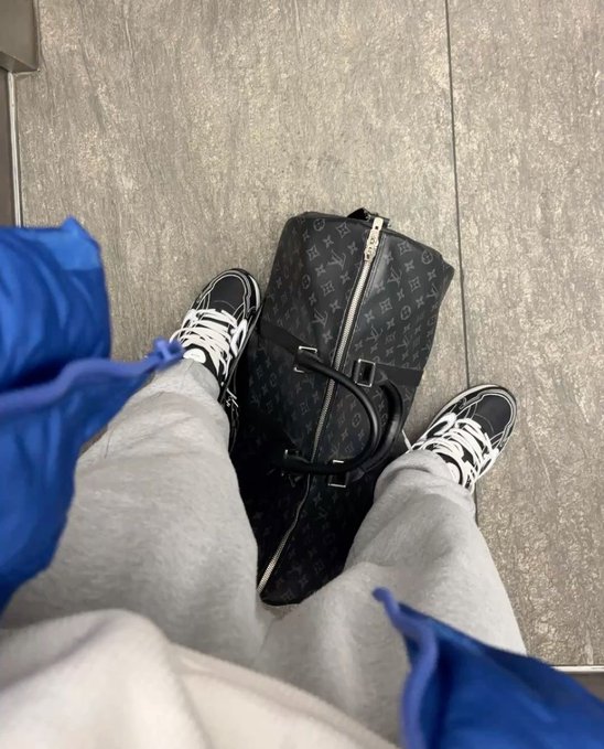 A first-person view looking down at someone's legs in gray sweatpants and black/white sneakers, with a black Louis Vuitton monogram duffel bag placed between their feet on a gray tiled floor.
