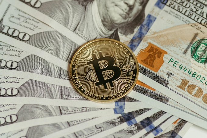A gold Bitcoin coin rests atop a fanned arrangement of U.S. one-hundred-dollar bills, with visible text including "100," "FRANKLIN," and "PE4444802." The image symbolizes the proposed $100M Bitcoin-backed municipal bond that New Hampshire's Executive Council rejected in a 3-2 vote.