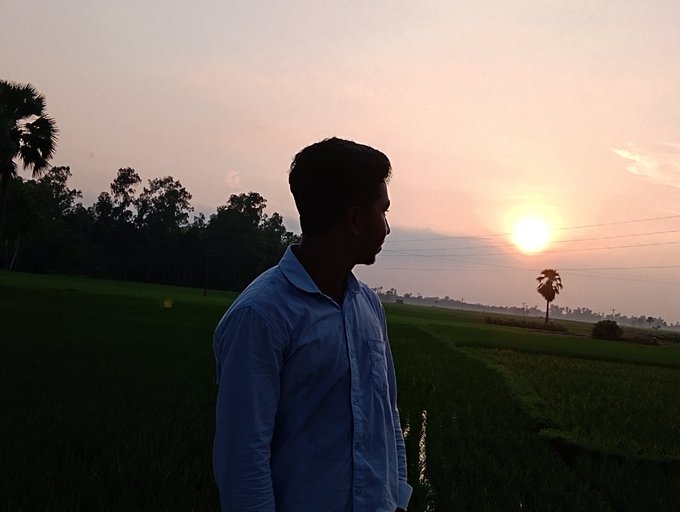 A young man in a light blue shirt stands in profile in a green field at sunset, with palm trees silhouetted against the warm orange and pink sky.