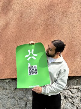 The image features a man holding a green poster with a white logo of a question mark. The poster has a QR code, which could be used for scanning or entering information into a website. The man appears to be posing with the poster against a brick wall backdrop.