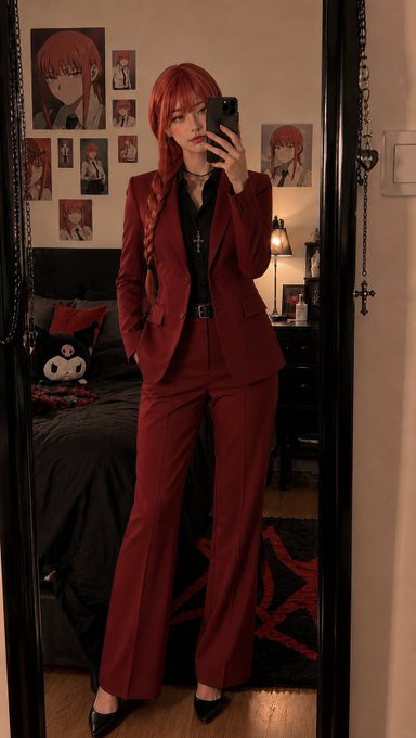 A mirror selfie of a person with long red braided hair wearing a fitted burgundy suit and black heels, standing in a bedroom decorated with anime character posters and a Kuromi plush.