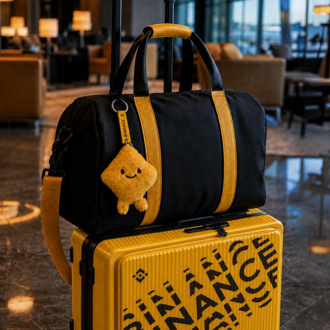 A black and yellow Binance-branded duffel bag sits atop a matching yellow hard-shell suitcase with 'BINANCE' printed on it, displayed in what appears to be a hotel lobby, with a small plush Binance mascot keychain attached.