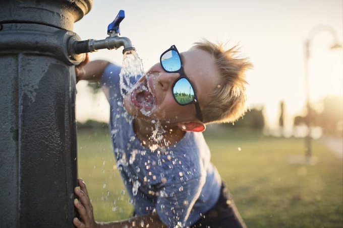 A young boy wearing reflective sunglasses tilts his head back with his mouth open as water splashes from a vintage outdoor metal fountain tap.