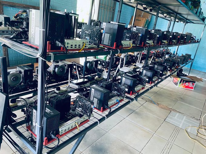 A photograph of multiple cryptocurrency mining rigs arranged on multi-tiered metal shelving units, each equipped with motherboards, GPUs, and power supplies.