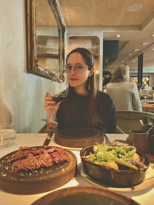 A woman wearing glasses holds a glass of red wine while seated at a restaurant table with dishes including steak and a skillet of vegetables in front of her.