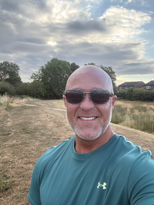 A bald, smiling man with a gray goatee wearing sunglasses and a teal Under Armour shirt takes a selfie in a grassy field with houses and trees in the background under a cloudy sky.