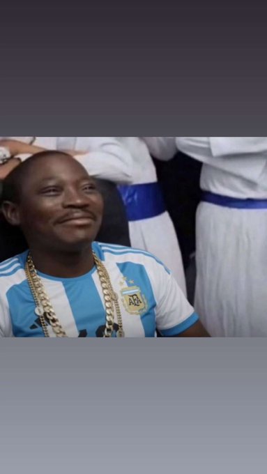 A man wearing an Argentina national football team jersey and gold chains, smiling slightly, with other people in white uniforms visible in the background.