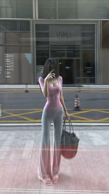 A woman takes a mirror selfie on a city sidewalk, wearing a pink short-sleeved top and light-colored pants while holding a large dark bag. Reflections of the street, traffic cones, and building entrances are visible in the glass behind her.