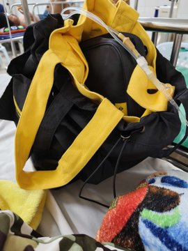 A black Binance tote bag with yellow straps sits open on a hospital bed, packed with essentials next to an IV pole. The branded bag, visible in its everyday utility, supports the post's message that good crypto swag transcends events by being a practical companion during personal moments like caring for a child in the hospital.