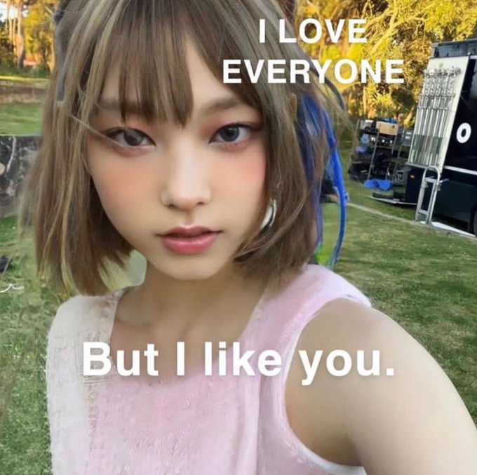 A young woman with short brown hair and blue streaks takes a selfie outdoors with text reading 'I LOVE EVERYONE' and 'But I like you.'