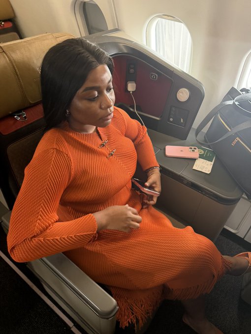 A woman in an orange ribbed knit dress sits in a premium airline cabin seat, focused on her phone while her passport and boarding documents rest on the side console beside her carry-on luggage.