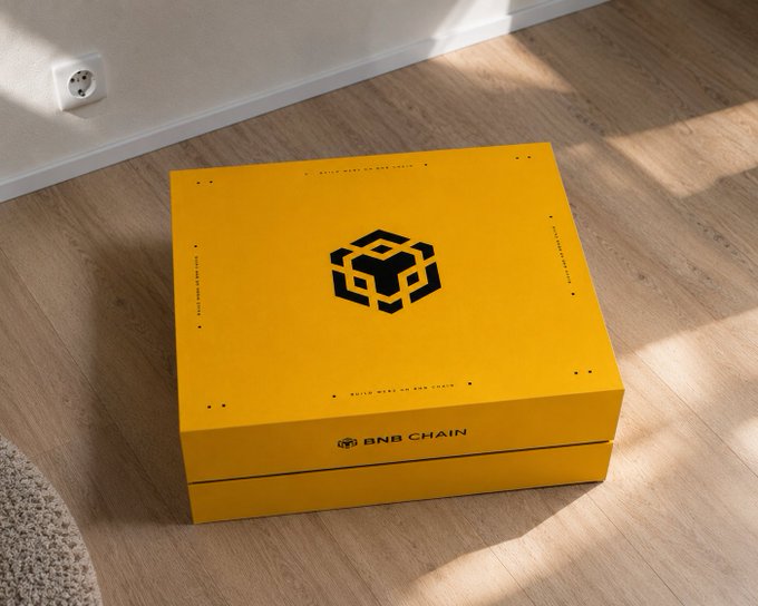 A yellow BNB Chain branded gift box sits on a wooden floor, featuring the distinctive black cube logo and "BNB CHAIN" text along the front edge. The unboxing setup reflects the excitement of receiving exclusive BNB Chain merchandise as mentioned in the post.