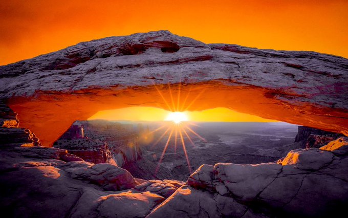 A vibrant sunrise bursts through a natural sandstone arch over a vast canyon landscape, its golden rays framed by rugged rock formations and a glowing orange sky.
