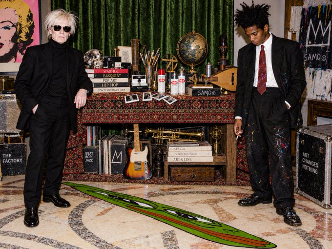 Two men in suits—one with white hair and sunglasses, the other holding a cigarette—pose in an art-filled room with a Basquiat-style painting of a face, a globe, spray paint cans labeled "SAMO," and visible texts including "WARHOL BASQUIAT POP ART INTERIOR CONTEMPORARY ART," "THE FACTORY," "ART CHANGES NOTHING," and "AFRICA The New York School ART & LIFE." The scene echoes the avant-garde, collaborative spirit of Warhol and Basquiat, aligning with the post's promotion of an art NFT collection on Objkt.