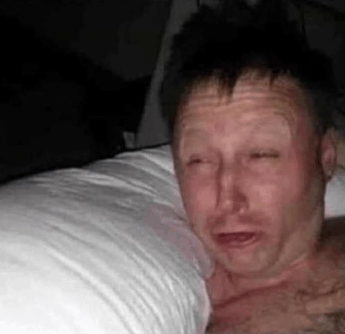 A close-up photo of a man lying in bed with a white pillow, appearing disheveled with a tired or distressed expression in low lighting.