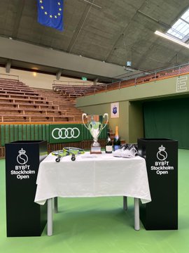 A silver championship cup and tennis rackets are displayed on a table covered with a white cloth, set on a green tennis court with "Bybit Stockholm Open" banners and an Audi logo in the background. The setup showcases the tournament's trophy presentation, directly connecting to the "Bybit Stockholm Open" branding as the event's sponsor.