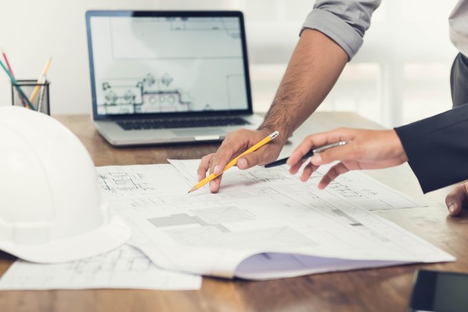 A close-up photo shows two people's hands pointing at architectural blueprints on a wooden desk, with a white hard hat, colored pencils, and a laptop displaying a floor plan in the background.