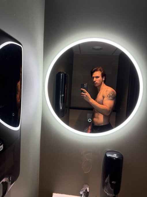 A shirtless man with earbuds takes a mirror selfie in a bathroom illuminated by a round LED mirror, holding a phone.