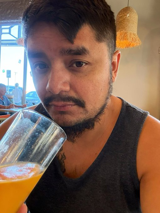 A close-up selfie of a bearded young man in a gray tank top holding a glass of amber-colored beverage, taken inside a venue with decorative hanging lamps.