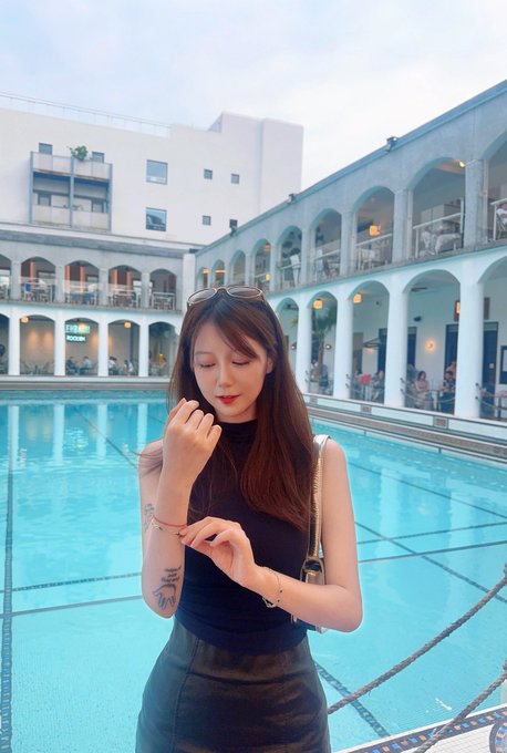 A young woman wearing a black top and skirt stands by a swimming pool in a courtyard, looking at her wrist as if checking a watch, with sunglasses perched on her head.