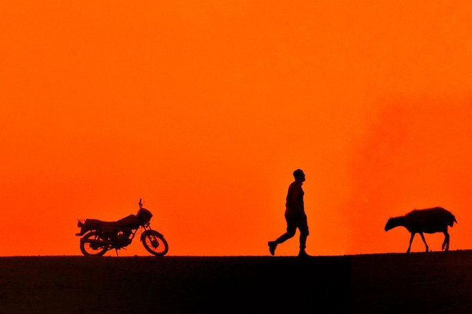 Silhouette of a man walking alongside a sheep with a motorcycle parked nearby against a vivid orange sky at sunset.