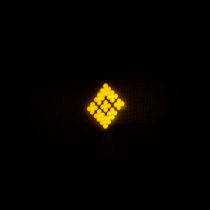 A glowing yellow LED sign in the shape of a Bitcoin "₿" symbol is displayed against a dark background, representing 24/7 cryptocurrency trading. The symbol visually reinforces the post's message about nonstop trading, with the Bitcoin logo serving as an iconic representation of the crypto market that never sleeps.
