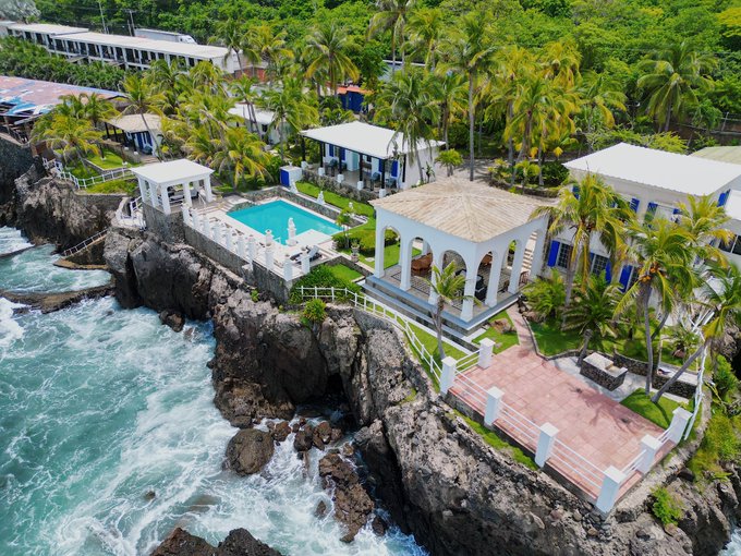 An aerial view of a luxurious oceanfront property featuring a pool, white pavilions, and a gazebo perched on rocky cliffs above crashing waves.