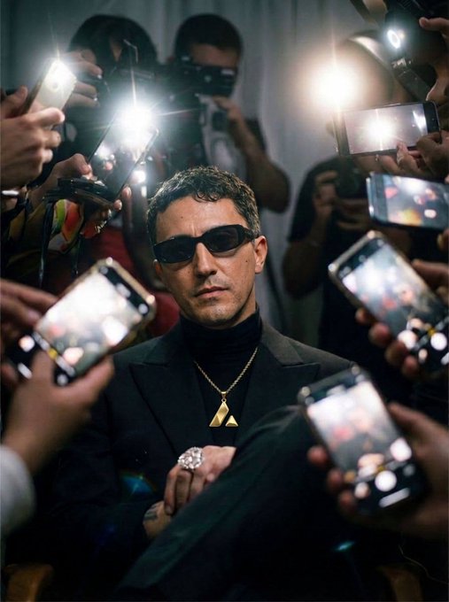 A man in a black suit, sunglasses, and gold triangular pendant sits confidently surrounded by a crowd of people photographing him with phones and cameras with bright flashes.