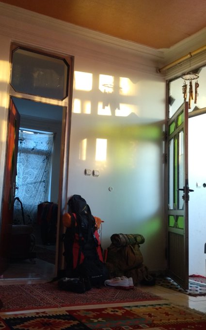 An interior entryway of a home with traditional patterned rugs, hiking backpacks and a rolled sleeping mat piled against a white wall, sunlight streaming through glass-paneled doors and a small window.